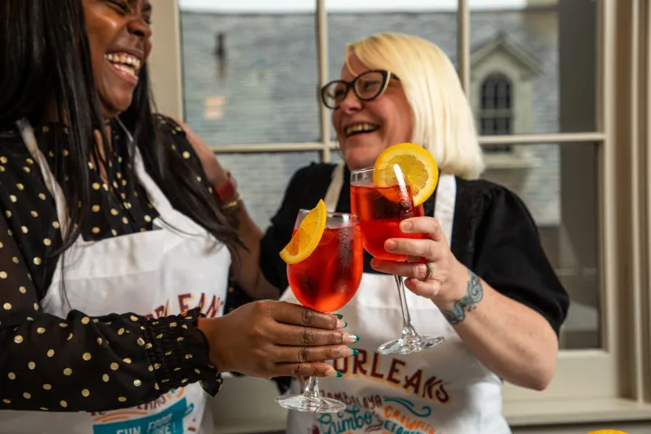 Two people in aprons laughing and clinking glasses with orange slices.