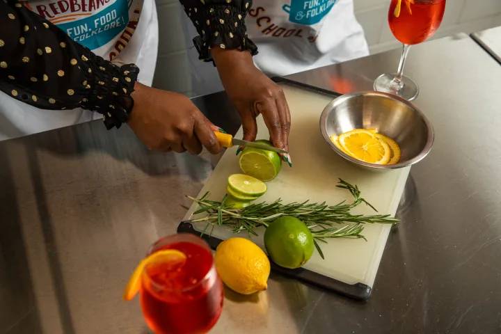 Person slicing lime on a cutting board with herbs, citrus, and two red drinks nearby.