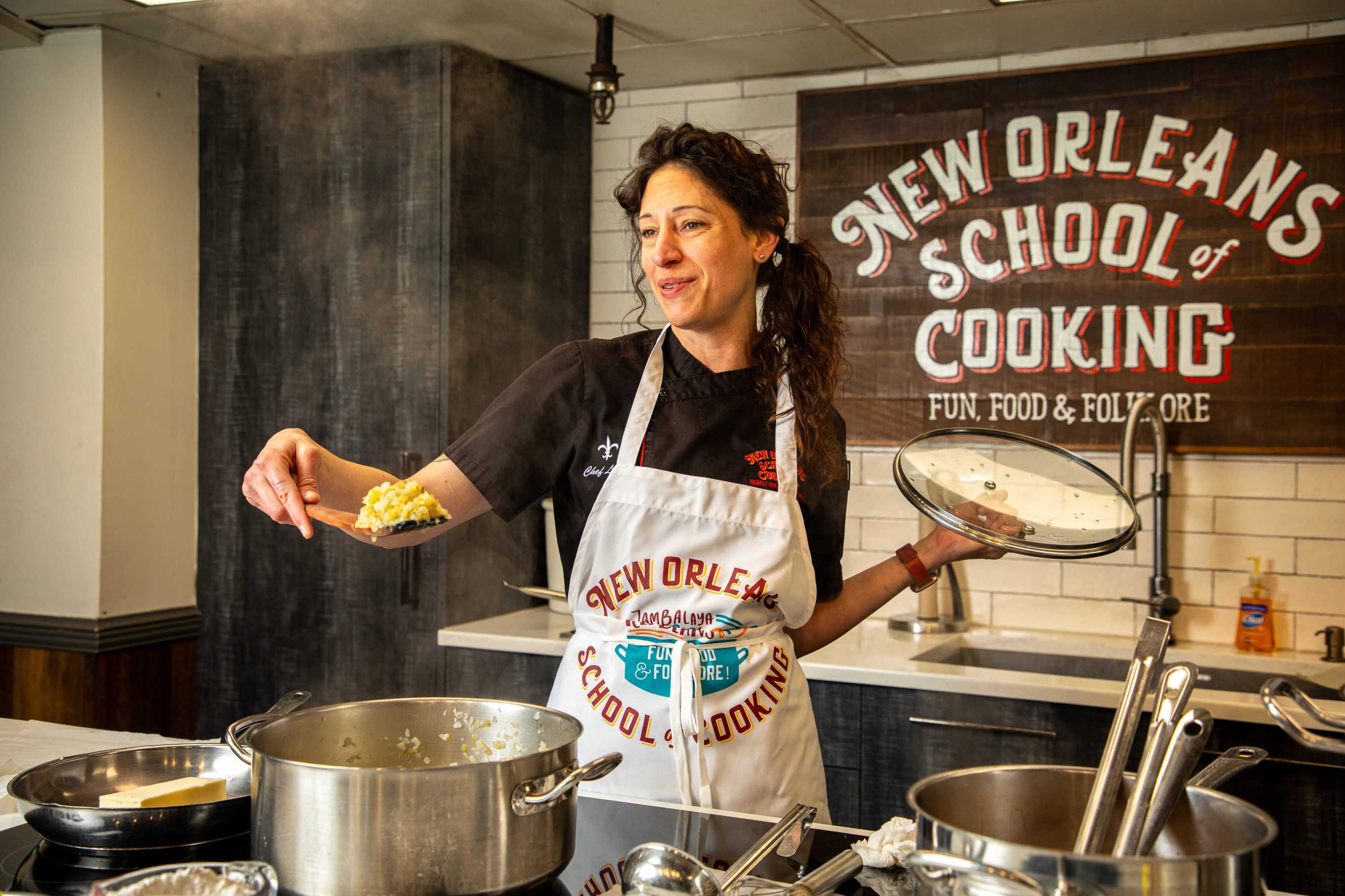 Chef cooking in kitchen at New Orleans School of Cooking, holding spoon and pot lid.