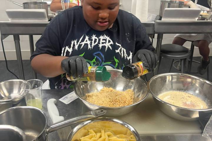 Child in cooking class preparing food with pasta and spices on table.