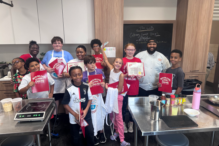 Group of kids and a chef holding certificates and cooking tools in a kitchen setting.
