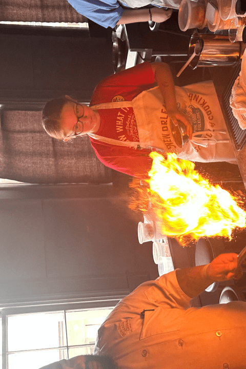 Chef performing a flambé technique in a kitchen with a large flame rising from the pan.