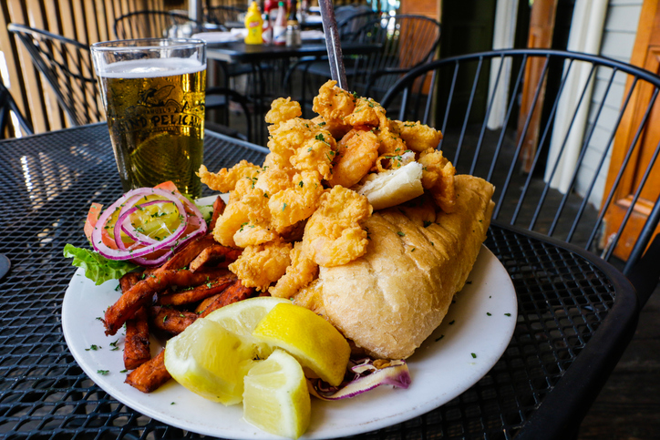 A crispy fried shrimp po’boy sandwich on French bread with lettuce and sauce, paired with golden sweet potato fries and a cold draft beer, represents the classic flavors of New Orleans cuisine.
